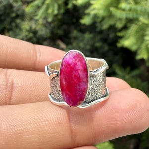 Puede incluir: Un anillo de plata con una gran piedra preciosa de color rosa magenta vibrante en forma ovalada en el centro. La banda tiene un acabado de plata mate texturizado ancho con bordes de plata lisos y elevados que enmarcan la piedra central.