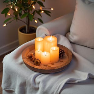 Könnte beinhalten: Ein Holztablett mit fünf brennenden, elfenbeinfarbenen Kerzen unterschiedlicher Höhe. Dazu getrocknete Blumen und ein Tannenzapfen. Das Tablett steht auf einer weißen Decke, die über einem Stuhl drapiert ist, mit einer kleinen Pflanze und Lichterketten im Hintergrund.
