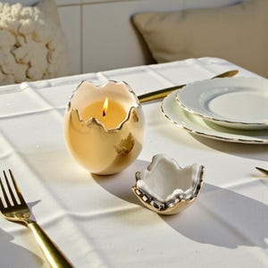 Może przedstawiać: Zapalone świeca w złotym, jajowatym świeczniku na białym obrusie. Mała, pasująca miseczka i złote sztućce również znajdują się na stole. Płomień świecy jasno się pali.