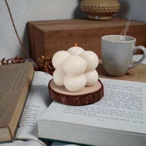 May include: A cream-colored bubble candle with a lit wick, resting on a wooden slice. The candle is surrounded by a book, a mug, and other decorative items. The candle's unique shape and warm glow create a cozy atmosphere.