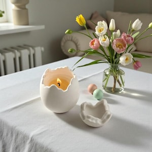 Może przedstawiać: Zapalone, białe świeca w kształcie jajka z postrzępioną krawędzią stoi na białym obrusie. W pobliżu znajduje się mała, pasująca miseczka w kształcie skorupki jajka. Wazon z różowymi, białymi i żółtymi tulipanami oraz pastelowe pisanki dopełniają scenę.