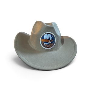 May include: A light beige cowboy hat featuring a circular patch on the front. The patch displays the text NY ISLANDERS in white and orange lettering, accompanied by a blue and orange graphic of a hockey stick and puck on a blue background.