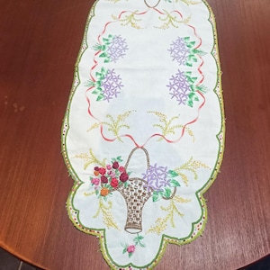 May include: Cream-colored embroidered table runner with a scalloped edge. Features floral basket designs with red, purple, and pink flowers, and green accents. Red ribbons and green trim complete the design.