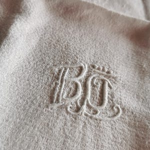 Pode incluir: Close-up de uma toalha bege clara com um monograma bordado. O monograma apresenta as letras "BC" em uma fonte cursiva decorativa. A toalha tem uma aparência tecida texturizada.