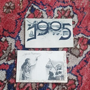 May include: Two antique postcards. The top one features the year "1905" with illustrations of buildings. The bottom postcard has illustrations of women and the years "1903" and "1904". Both postcards are on a red and patterned surface.