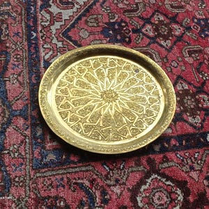 Op de afbeelding: Een ronde, goudkleurige metalen schaal met een ingewikkeld geometrisch sterpatroon in het midden. De schaal staat op een rood en blauw patroonkleed, wat een contrast van texturen en kleuren creëert. Het ontwerp van de schaal suggereert een decoratief of dienend doel.