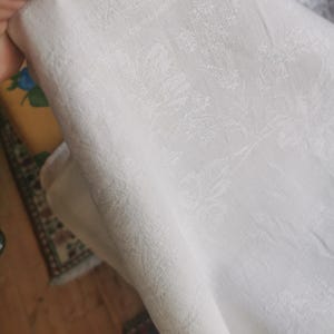 Pode incluir: Close-up de um tecido branco com um padrão floral sutil. O tecido está dobrado, revelando sua textura e design. O fundo está desfocado, focando a atenção no têxtil detalhado.