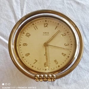 May include: A vintage gold-toned desk clock with a cream-colored face. The clock features the brand name "OPIS" and the text "15 JEWELS". The clock has a circular design with a gold-toned ring and a decorative base. The clock measures approximately 3 inches in diameter.