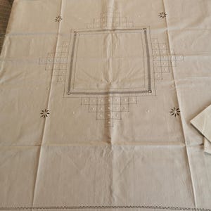 Pode incluir: Toalha de mesa creme com padrões geométricos e florais bordados. O design central apresenta uma moldura quadrada com uma borda pontilhada, rodeada por padrões de diamantes e quadrados. Motivos florais são colocados nas bordas.