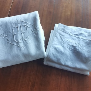 May include: Two folded white linen pillowcases with embroidered initials and floral accents. The initials "PP" are centered within a diamond-shaped design. The pillowcases are neatly folded on a wooden surface.