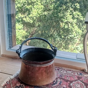 May include: A copper cauldron with a black handle and rim sits on a patterned rug. The cauldron has a wide base and tapers upwards. The background shows a window with a view of green trees.