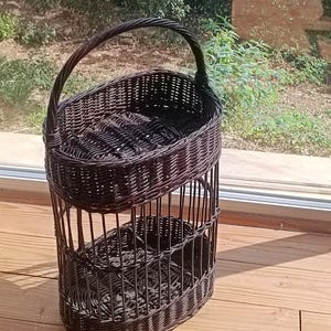 May include: A dark brown, two-tiered wicker basket with a handle. The basket has an oval shape and is made of woven wicker. The handle is arched and attached to the top tier. The basket is sitting on a wooden surface.