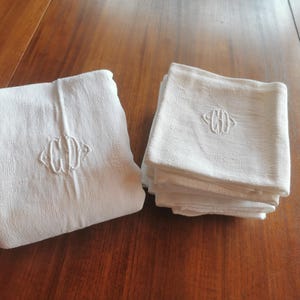 Pode incluir: Duas toalhas de mesa de linho branco com monogramas bordados. Uma toalha de mesa está desdobrada e a outra está dobrada em uma pilha. As toalhas de mesa estão em uma superfície de madeira. O monograma parece ser um "W" e "H" estilizados.