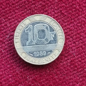 以下が含まれることがあります： 10と1989の数字が入った銀と金のコイン。コインには「LIBERTE」、「EGALITE」、「FRATERNITE」の文字が刻まれています。赤い表面に置かれています。
