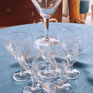 Könnte beinhalten: Eine Sammlung klarer, geätzter Weingläser, die pyramidenförmig angeordnet sind. Jedes Glas hat ein Blumendesign. Die Gläser stehen auf einer blauen Tischdecke und erzeugen eine klassische und elegante Ästhetik.