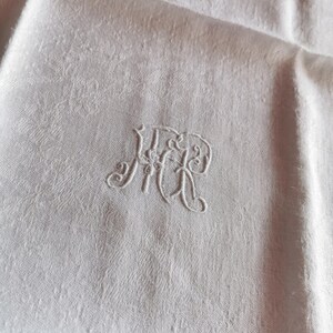 Pode incluir: Close-up de um guardanapo de linho branco com um monograma bordado. O monograma apresenta as letras "JHR" em uma elegante escrita cursiva. O tecido tem um padrão floral sutil.