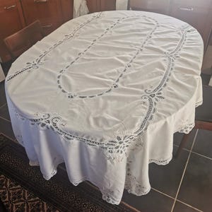 Pode incluir: Toalha de mesa oval branca com detalhes intrincados de renda nas bordas e ao longo do design central. A toalha de mesa é drapeada sobre uma mesa de madeira, mostrando seu design elegante e textura. A toalha de mesa é feita de tecido branco.