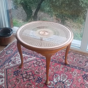 May include: A round, wooden side table with a woven cane top and three curved legs. The table is approximately 61 cm in diameter and stands about 51 cm tall. The table is set on a patterned rug.