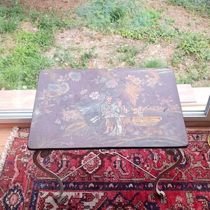 May include: A rectangular antique table with a dark, ornate top featuring a painted scene of figures and floral elements. The table rests on a wrought iron base, set atop a red patterned rug.