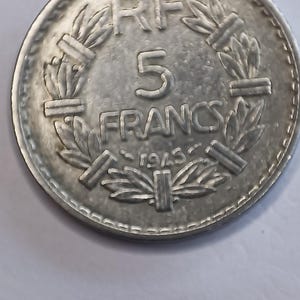 May include: A silver 5 Francs coin from 1945, featuring the text "RF" and "FRANCS" with the number "5" in the centre. The coin is encircled by a laurel wreath design.
