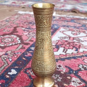 Könnte beinhalten: Eine Messingvase mit aufwendigen, geätzten Blumen- und geometrischen Mustern. Die Vase hat einen ausgestellten oberen Rand, einen schlanken Körper und einen runden Sockel. Der Hintergrund ist ein farbenfroher Teppich. Der Text "FRENCH ANTIKS" ist sichtbar.