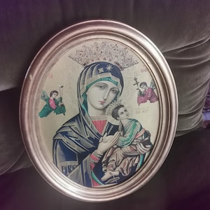 Puede incluir: Icono religioso enmarcado ovalado que representa a la Virgen María sosteniendo a un niño, con dos ángeles a cada lado. El icono presenta colores dorados, azules, rojos y verdes. El marco es de color dorado.