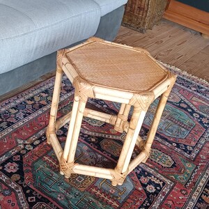 May include: A small, octagonal bamboo side table with a woven top. The table is light brown and sits on a patterned rug with red, blue, and green hues. The table is approximately 18 inches tall.