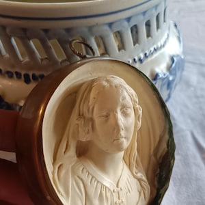 May include: A vintage cameo pendant with a carved relief of a woman's face in ivory tones, set in a bronze-coloured frame. A blue and white ceramic decorative piece is in the background.