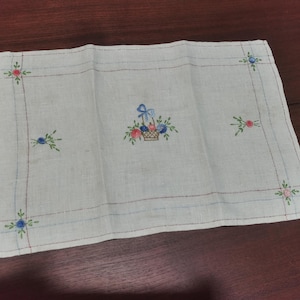 May include: A white linen handkerchief with embroidered floral details. The center features a basket of flowers with a blue bow. The corners and edges are adorned with small floral sprigs in red, blue, and pink.