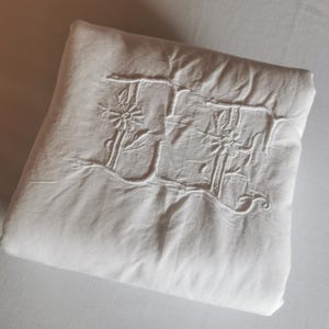 May include: A folded white linen sheet with embroidered floral details and the letter "J". The embroidery is in a matching white thread, creating a subtle, elegant design. The sheet is neatly folded, suggesting it is new or freshly laundered.