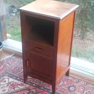 May include: A wooden nightstand with a drawer and a cabinet door. The nightstand is a warm brown color and has a light-colored top. It is placed on a patterned rug. The text "XIAOMI 11T | FRENCH ANTIKS" is visible at the bottom.