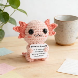 May include: A crocheted pink axolotl figurine with coral-colored gills and black eyes. The axolotl holds a sign that reads "Positiver Axolotl" in German, with an inspirational message. A mug and plant are in the background.