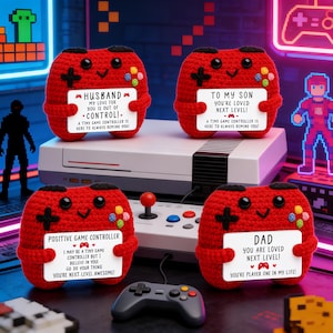 May include: Four red crocheted game controller plushies with black cross and colorful button details. Each controller holds a white sign with a different message, such as "Husband" or "Dad". A gray game controller sits in front of a retro game console.