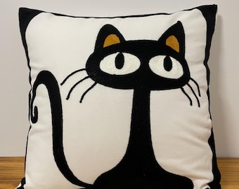 Puede incluir: Cojín cuadrado blanco con un diseño de gato negro. El gato está bordado en negro con ojos blancos grandes, bigotes largos y orejas internas doradas. El cojín tiene un borde negro.