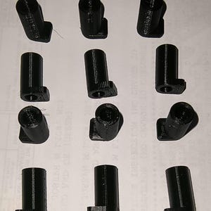 May include: A collection of twelve black, cylindrical plastic parts with a right-angle base. Each part has a hollow, open top and a small, rectangular base. The parts are arranged in a grid pattern on a light-colored surface.