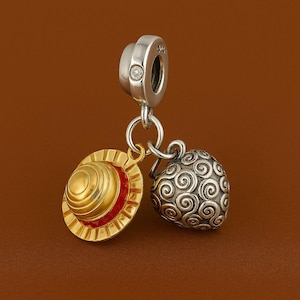 May include: A silver charm with a small diamond, connected to a gold and red hat charm and a silver, swirled design charm. The charms are suspended against a brown background.