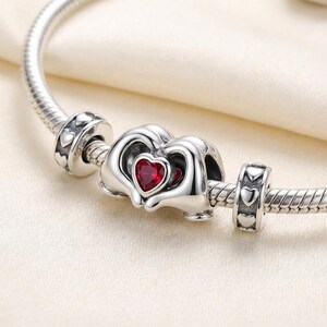 May include: A silver charm bracelet featuring heart-shaped beads. The central charm has a red heart-shaped stone set within a silver heart design. The bracelet is displayed against a white backdrop.