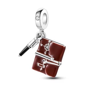 Puede incluir: Un charm de plata con un libro de esmalte marrón, un bolígrafo negro y un diseño de brújula. El charm tiene una pequeña gema transparente y está diseñado para llevarse en una pulsera o collar.