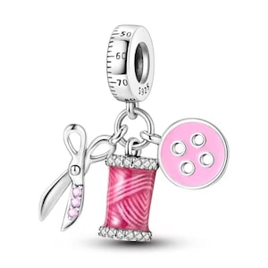Puede incluir: Un charm de plata con elementos de costura. Presenta un carrete de hilo rosa, un botón rosa y tijeras con gemas rosas. La parte superior tiene un anillo plateado con una escala de medición.