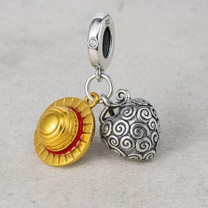 May include: A silver charm with a small diamond and two pendants. One pendant is a gold and red hat, and the other is a silver fruit with a swirl design. The charm is likely for a bracelet or necklace.