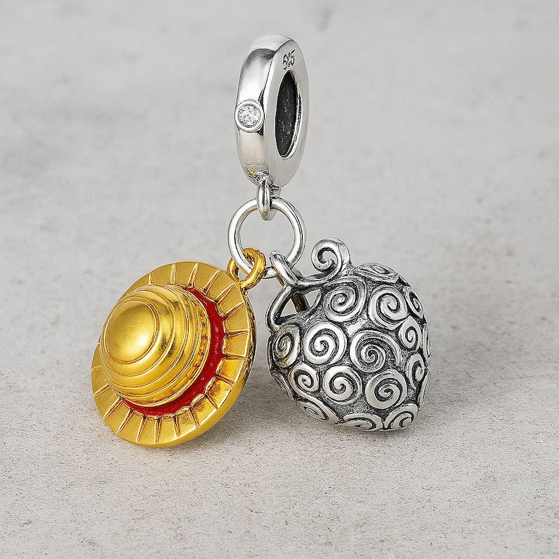 May include: A silver charm with a small diamond and two pendants. One pendant is a gold and red hat, and the other is a silver fruit with a swirl design. The charm is likely for a bracelet or necklace.