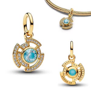 Può includere: Ciondolo dorato con una gemma blu chiaro al centro, circondata da cristalli trasparenti. Il ciondolo ha un design circolare con un motivo decorativo e un anello per attaccarlo a una catena o un braccialetto. Il testo "ARMOR UP" è visibile.