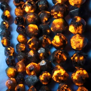 Könnte beinhalten: Eine Sammlung leuchtender, dunkel gefärbter Steine mit hellorangefarbener und gelber Innenbeleuchtung. Die Steine haben unregelmäßige Formen und unterschiedliche Größen, wodurch eine visuell auffällige Darstellung entsteht. Der Gesamteffekt erinnert an Miniatur-Leuchtkugeln.
