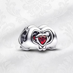 May include: A silver charm featuring a heart-shaped red gemstone. The charm has a layered design with heart-shaped cutouts. The background is a white, patterned surface.