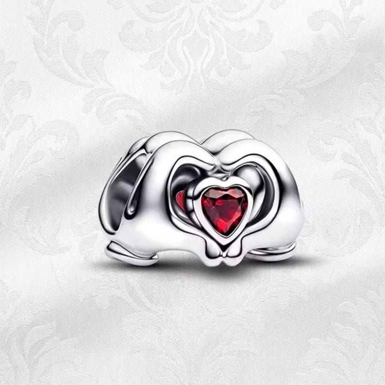 May include: A silver charm featuring a heart-shaped red gemstone. The charm has a layered design with heart-shaped cutouts. The background is a white, patterned surface.