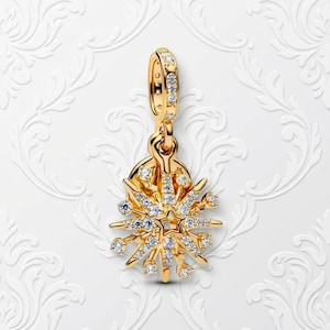 Puede incluir: Un colgante dorado con un diseño de estrella. El colgante está adornado con numerosas piedras preciosas claras y brillantes. El lazo superior y las puntas de la estrella también están adornados con piedras preciosas más pequeñas. El colgante cuelga sobre un fondo blanco estampado.