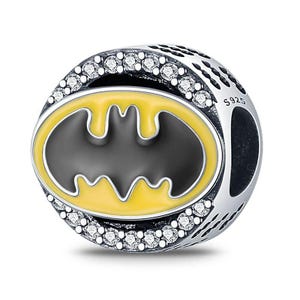 May include: A silver charm with a black and yellow Batman logo. The charm is round with a ring of small, clear stones around the logo. The charm is marked with "S925".