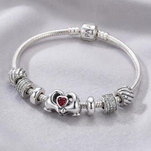 May include: A silver bracelet with a snake chain. It features charms, including one with "PAA" and a red heart charm. Other decorative beads are also present. The bracelet is displayed on a grey surface.