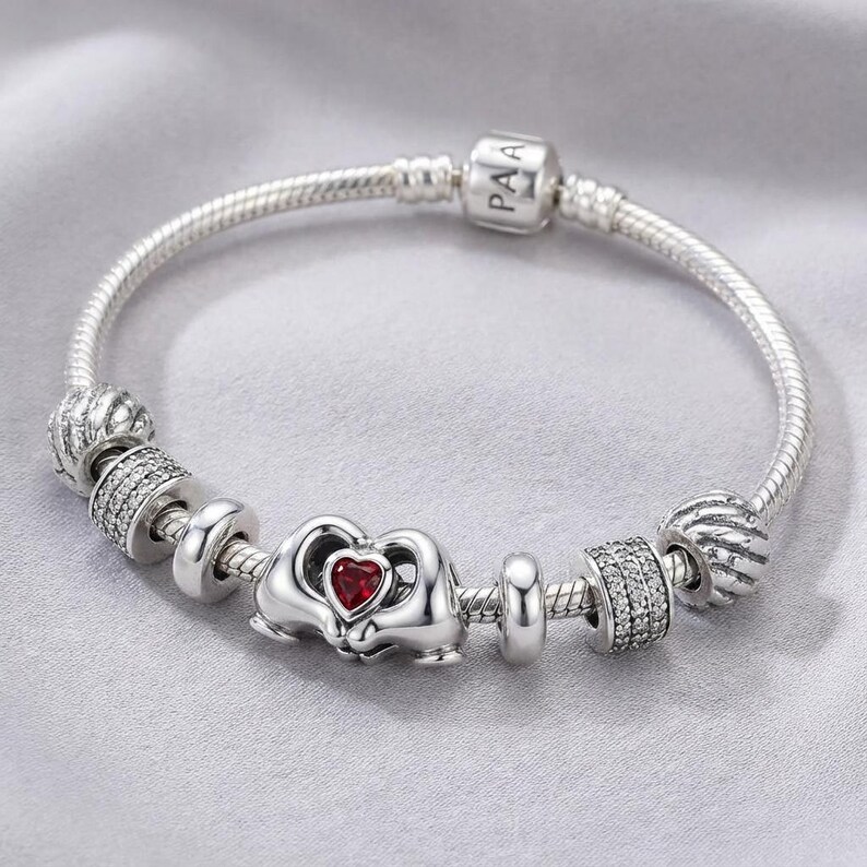 May include: A silver bracelet with a snake chain. It features charms, including one with "PAA" and a red heart charm. Other decorative beads are also present. The bracelet is displayed on a grey surface.