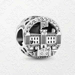 May include: A silver charm featuring a detailed depiction of a building with a Union Jack flag, likely Buckingham Palace. The charm is adorned with clear crystals and the word "LONDON" is engraved on the side. The design is intricate and the charm has a polished finish.